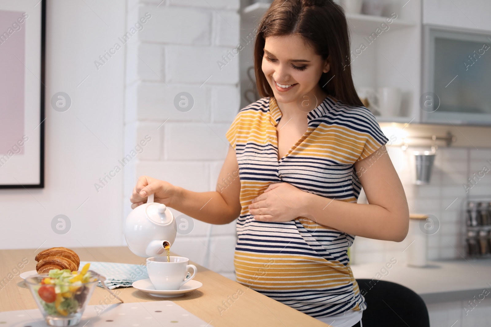 Photo of Young pregnant woman pouring tea into cup at table in kitchen