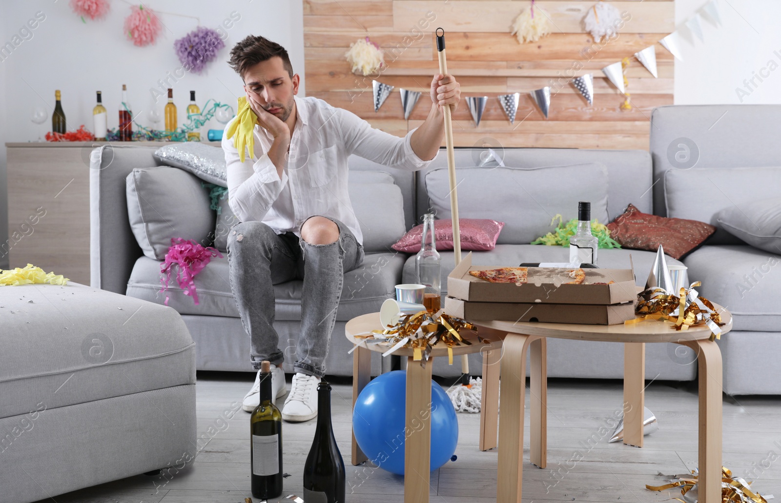 Photo of Young man with mop suffering from hangover in messy room after party