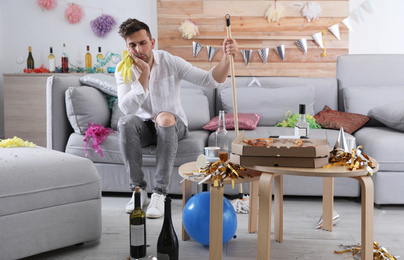 Young man with mop suffering from hangover in messy room after party