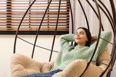 Photo of Young woman relaxing in hanging chair near window at home