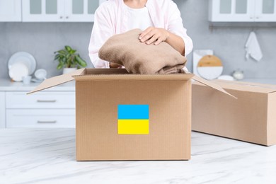 Photo of Humanitarian aid for Ukraine. Woman putting clothes in donation box at white table indoors, closeup