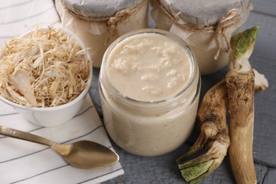 Photo of Spicy horseradish sauce in jars, roots and spoon on grey wooden table, closeup