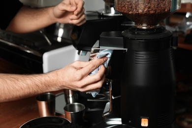 Photo of Barista cleaning coffee grinding machine with rag in cafe, closeup