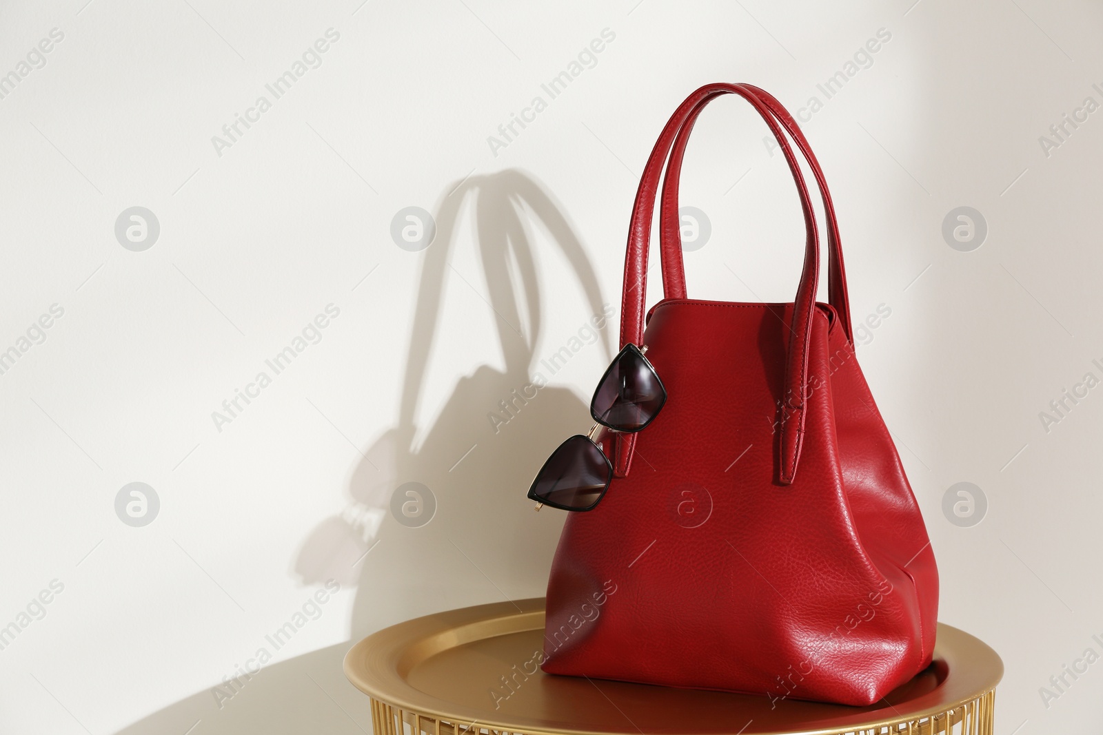 Photo of Stylish red woman's bag and sunglasses on table near light wall. Space for text