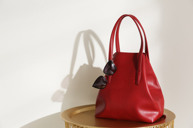 Stylish red woman's bag and sunglasses on table near light wall. Space for text