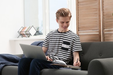 Photo of Online learning. Smiling teenage boy with laptop writing in notebook on sofa at home
