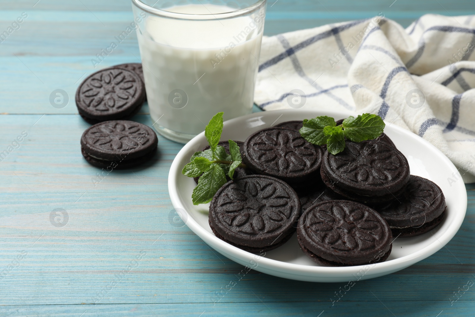 Photo of Plate with tasty sandwich cookies, mint and glass of milk on light blue wooden table, closeup