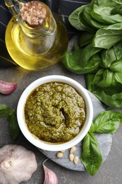 Photo of Tasty pesto sauce in bowl, basil, pine nuts, garlic and oil on grey table, top view