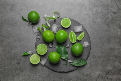 Photo of Slate plate with fresh ripe limes and ice cubes on gray background, top view