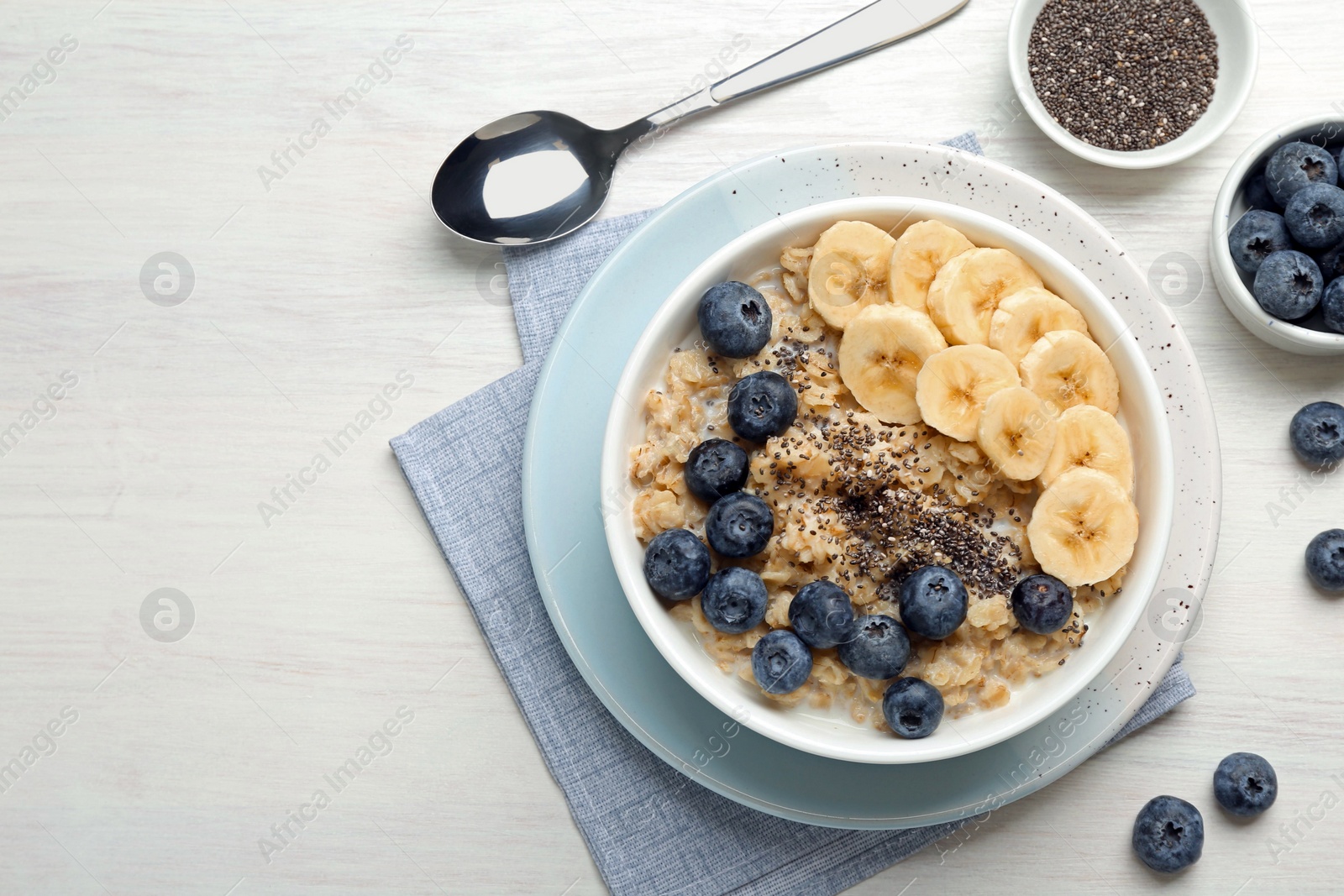 Photo of Tasty oatmeal with banana, blueberries and chia seeds served in bowl on white wooden table, flat lay. Space for text