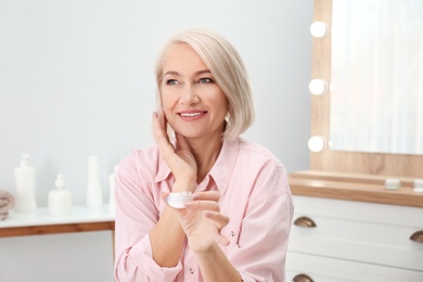 Portrait of charming mature woman with healthy beautiful face skin and natural makeup applying cream indoors