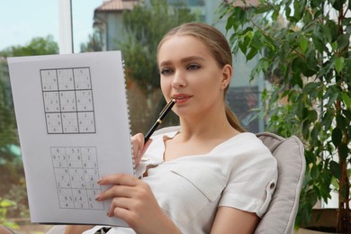 Photo of Beautiful young woman solving sudoku puzzle near window indoors