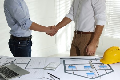 Architects shaking hands near table with construction projects in office, closeup