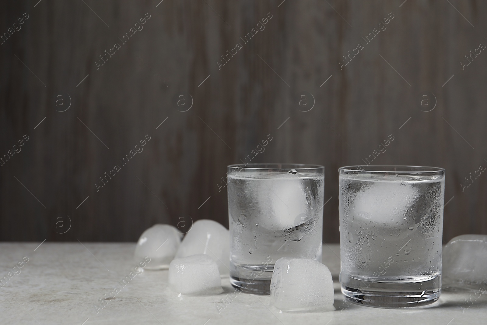 Photo of Shot glasses of vodka with ice cubes on light grey table against wooden background. Space for text