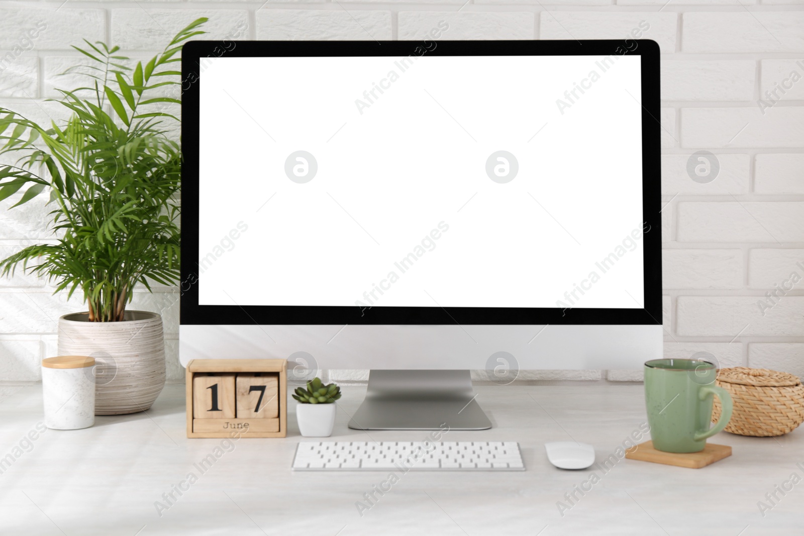 Photo of Office workplace with computer, glasses, cup, wooden block calendar and houseplant on light table near white brick wall