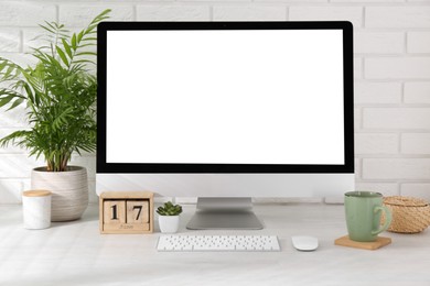 Photo of Office workplace with computer, glasses, cup, wooden block calendar and houseplant on light table near white brick wall