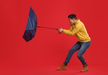 Emotional man with umbrella caught in gust of wind on red background