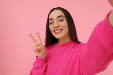 Photo of Smiling young woman taking selfie and showing peace sign on pink background