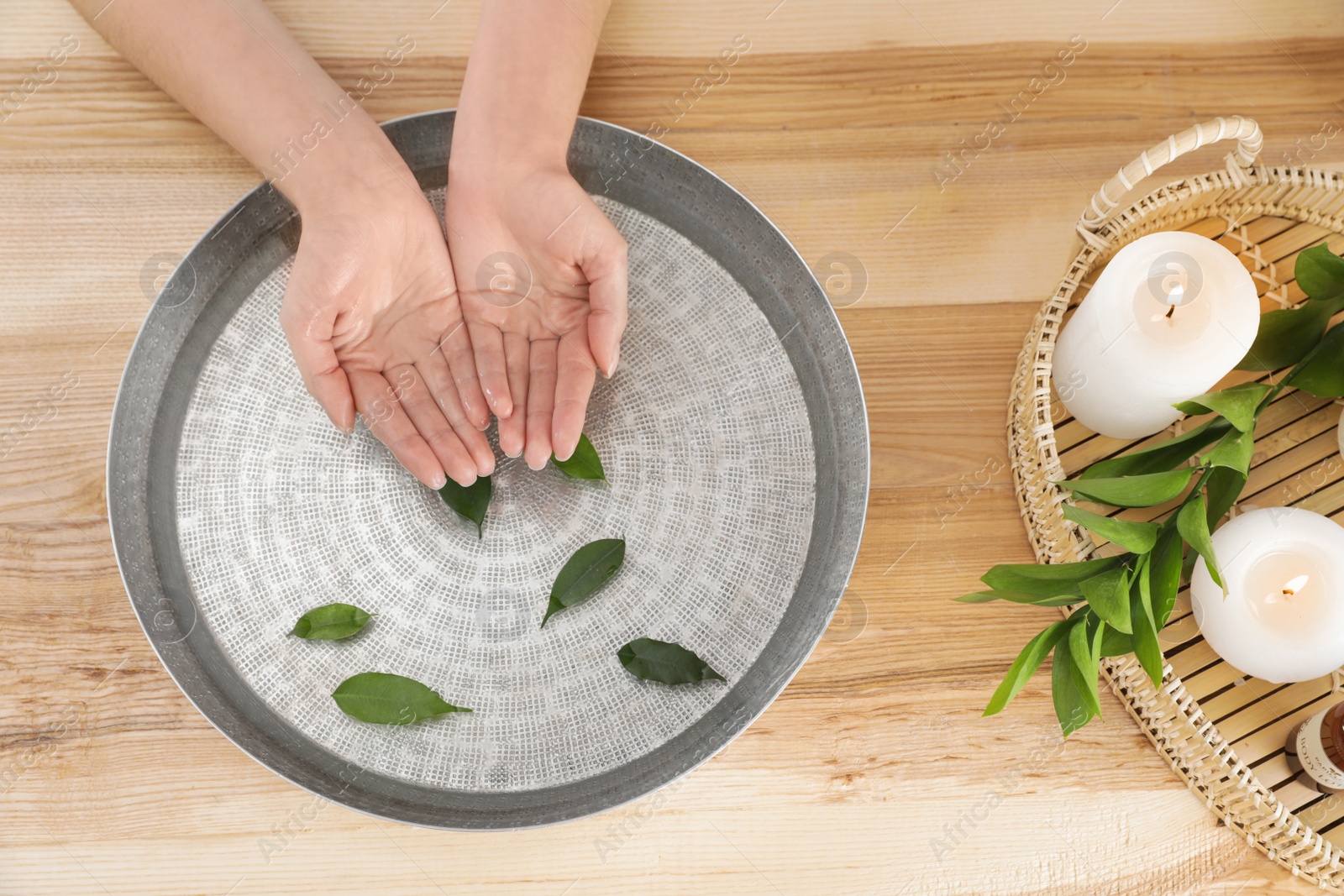 Photo of Woman soaking her hands in bowl with water and leaves on wooden table, top view. Spa treatment