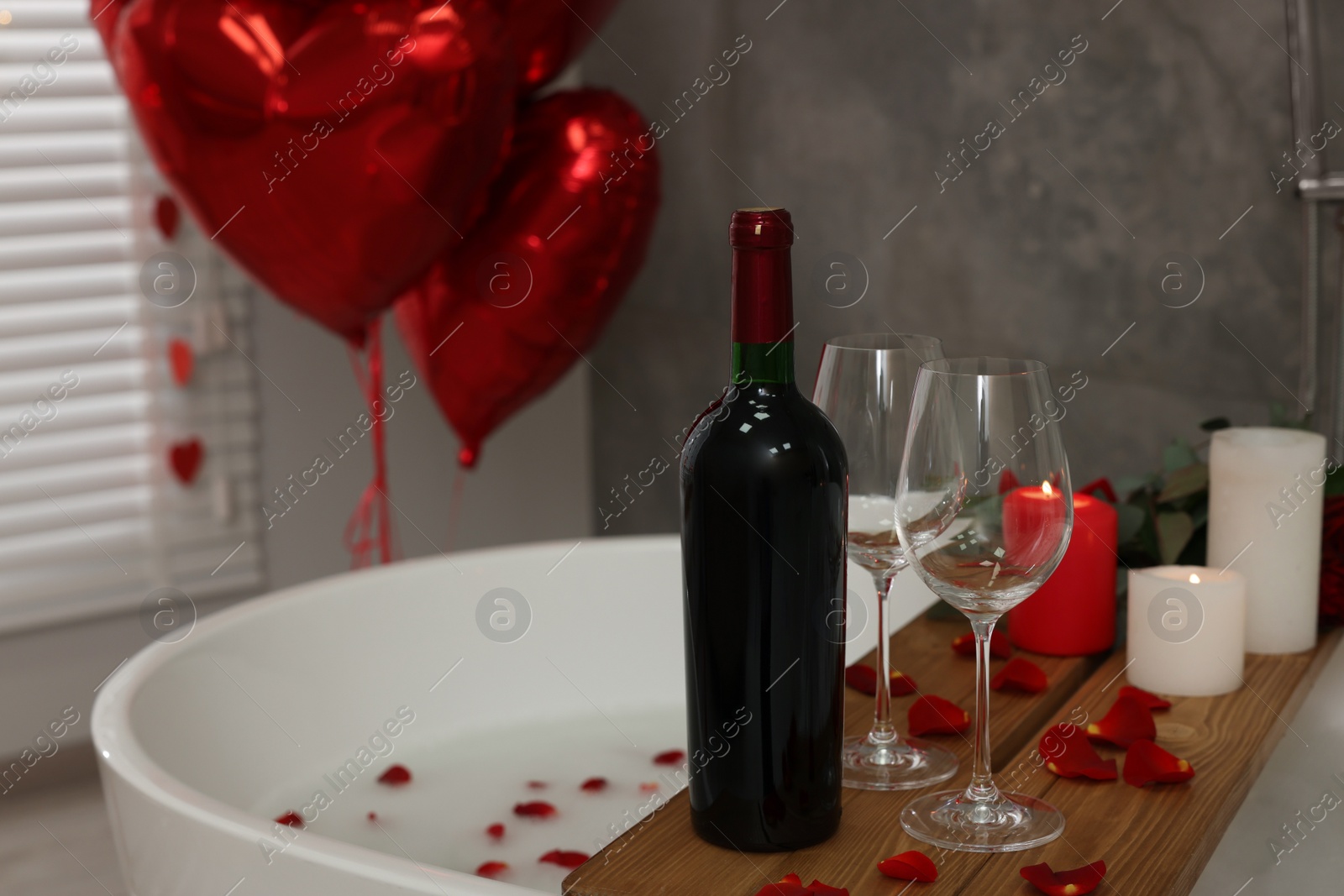 Photo of Wooden tray with wine, burning candles and rose petals on tub in bathroom. Valentine's day celebration