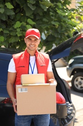 Young courier holding parcels near delivery car outdoors
