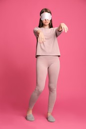 Photo of Young woman wearing pajamas, mask and slippers in sleepwalking state on pink background