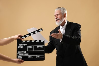 Photo of Senior actor performing role while second assistant camera holding clapperboard on beige background. Film industry