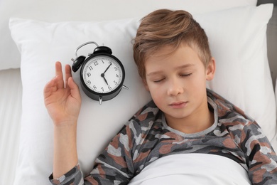 Cute boy with alarm clock sleeping in bed, above view