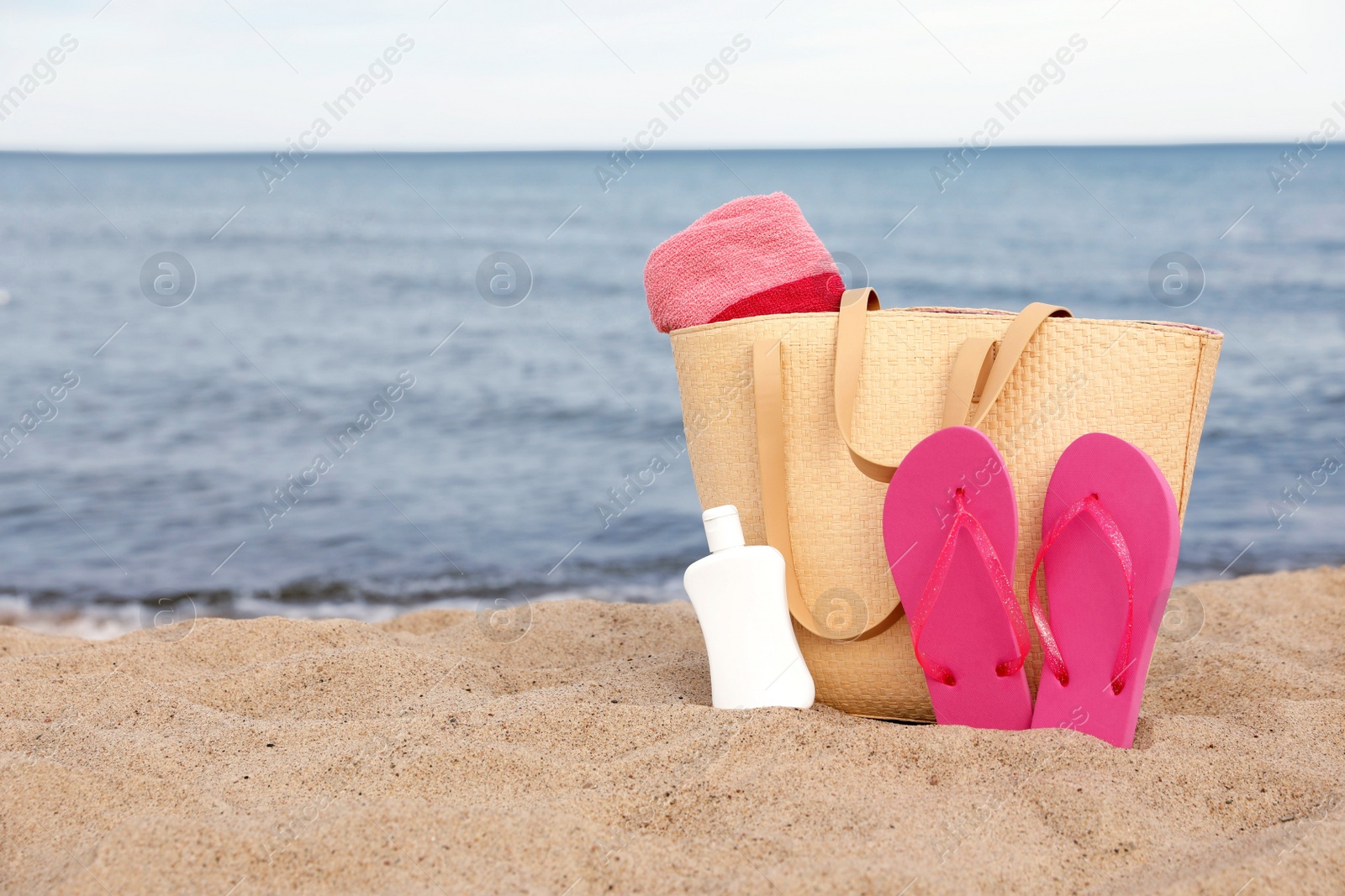 Photo of Summer bag with slippers, beach towel and sunscreen on sand near sea, space for text