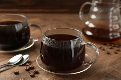 Photo of Hot coffee in glass cups, spoons and beans on wooden table, closeup