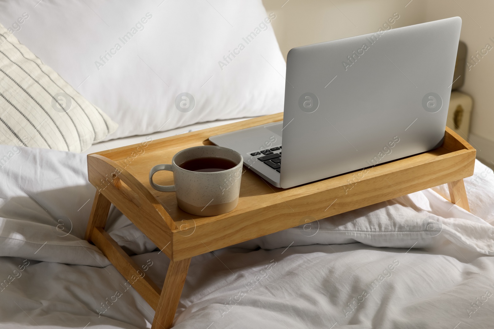 Photo of Wooden tray with modern laptop and cup of aromatic tea on bed indoors