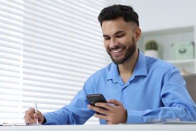 Happy young man using smartphone while working at white table in office, low angle view