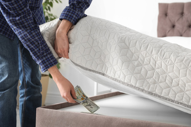 Photo of Man hiding dollar banknotes under mattress in bedroom, closeup. Money savings