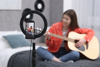 Photo of Teenage blogger playing guitar while streaming at home, focus on smartphone