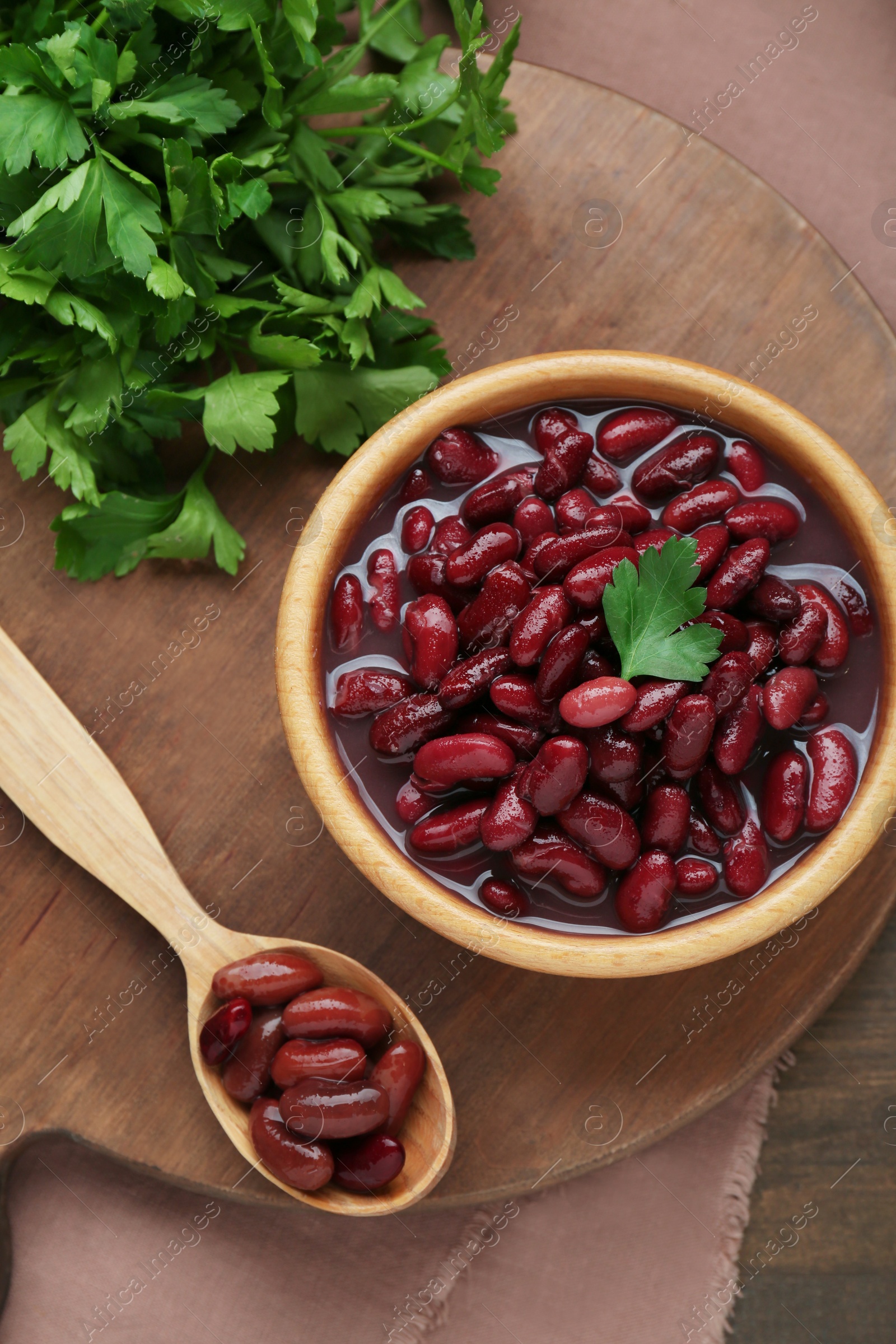 Photo of Bowl of canned red kidney beans, parsley and spoon on wooden table, flat lay