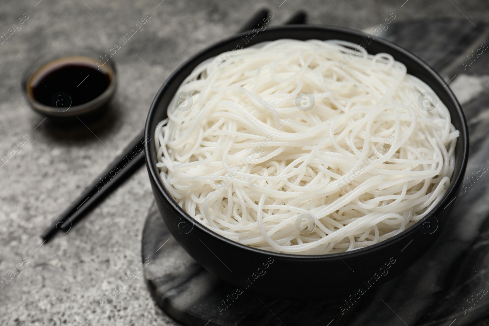 Photo of Bowl with cooked rice noodles, soy sauce and chopsticks on grey table, closeup