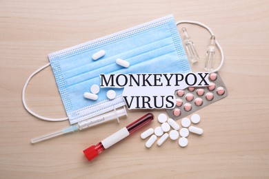 Photo of Card with words Monkeypox Virus, medical mask, different pills, syringe and test tube on wooden table, flat lay
