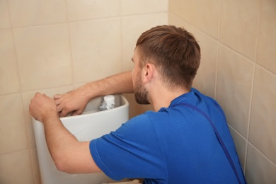 Professional plumber in uniform fixing toilet tank indoors