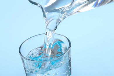 Photo of Pouring water from jug into glass on light blue background, closeup