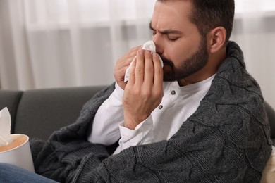 Photo of Sick man wrapped in blanket with tissue blowing nose on sofa indoors. Cold symptoms