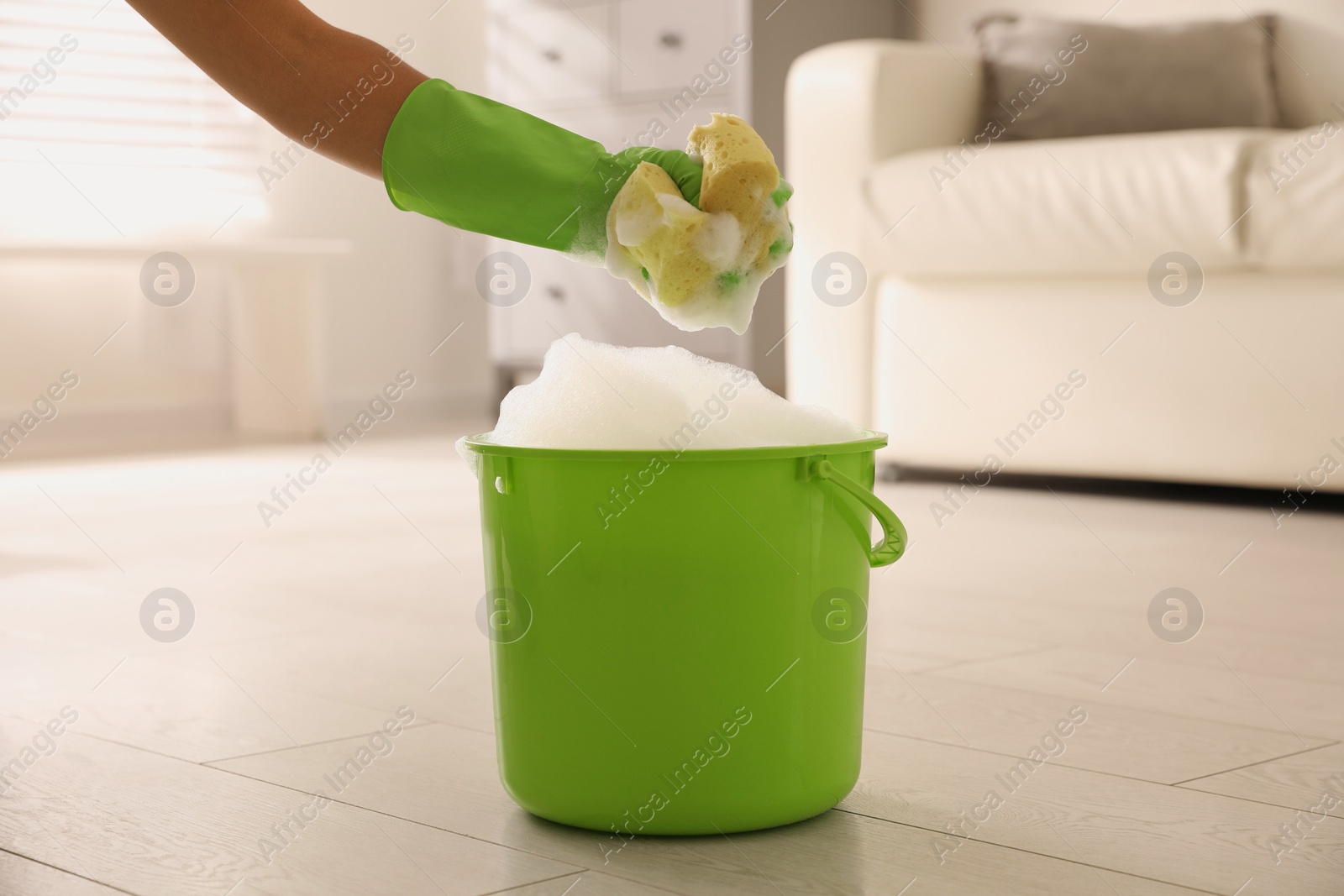 Photo of Woman holding sponge with foam over bucket indoors, closeup. Cleaning supplies