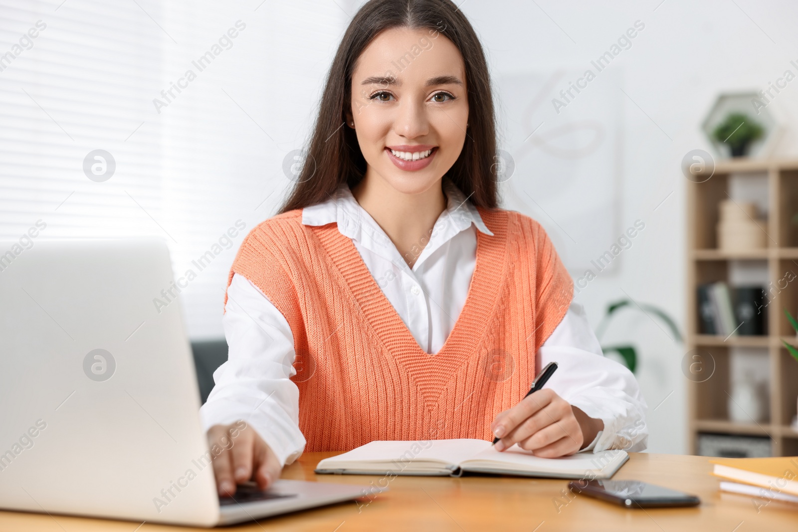 Photo of Young woman writing in notebook while working on laptop at wooden table indoors