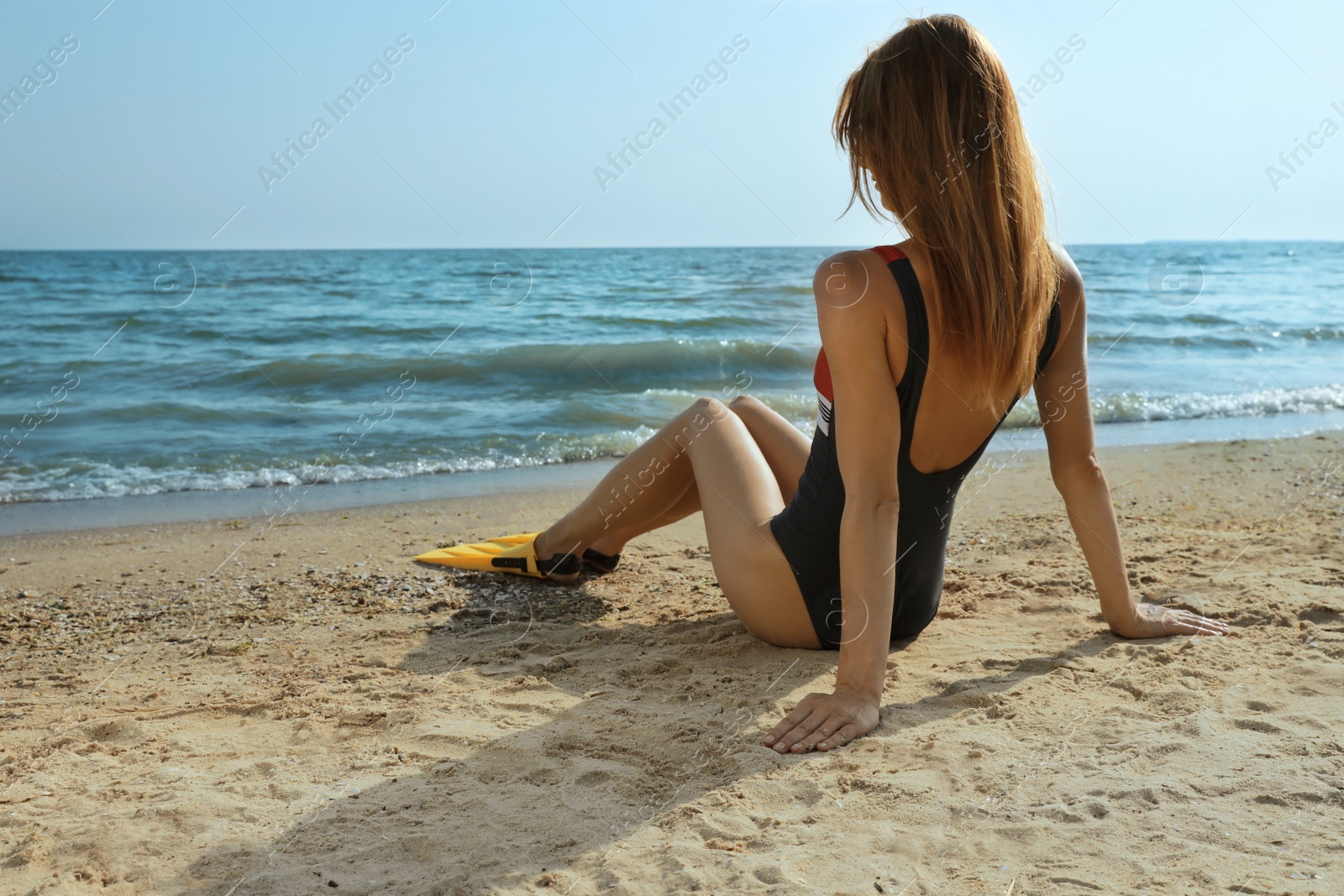 Photo of Woman in flippers sitting near sea on beach, back view