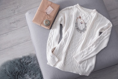 Photo of Cozy knitted sweater with jewelry and gift on pouf in living room, flat lay. Space for text