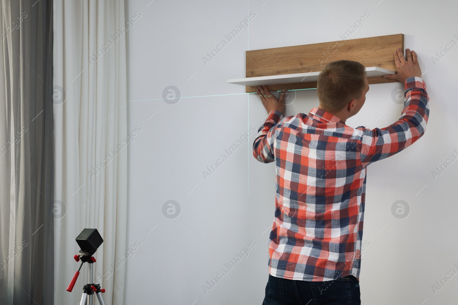 Photo of Man using cross line laser level for hanging wooden shelf on light wall indoors, back view