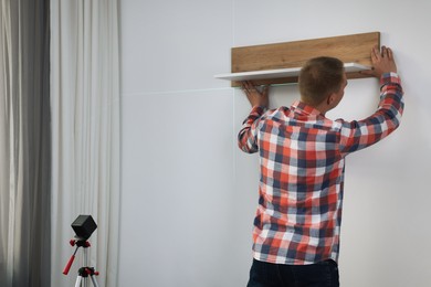 Man using cross line laser level for hanging wooden shelf on light wall indoors, back view