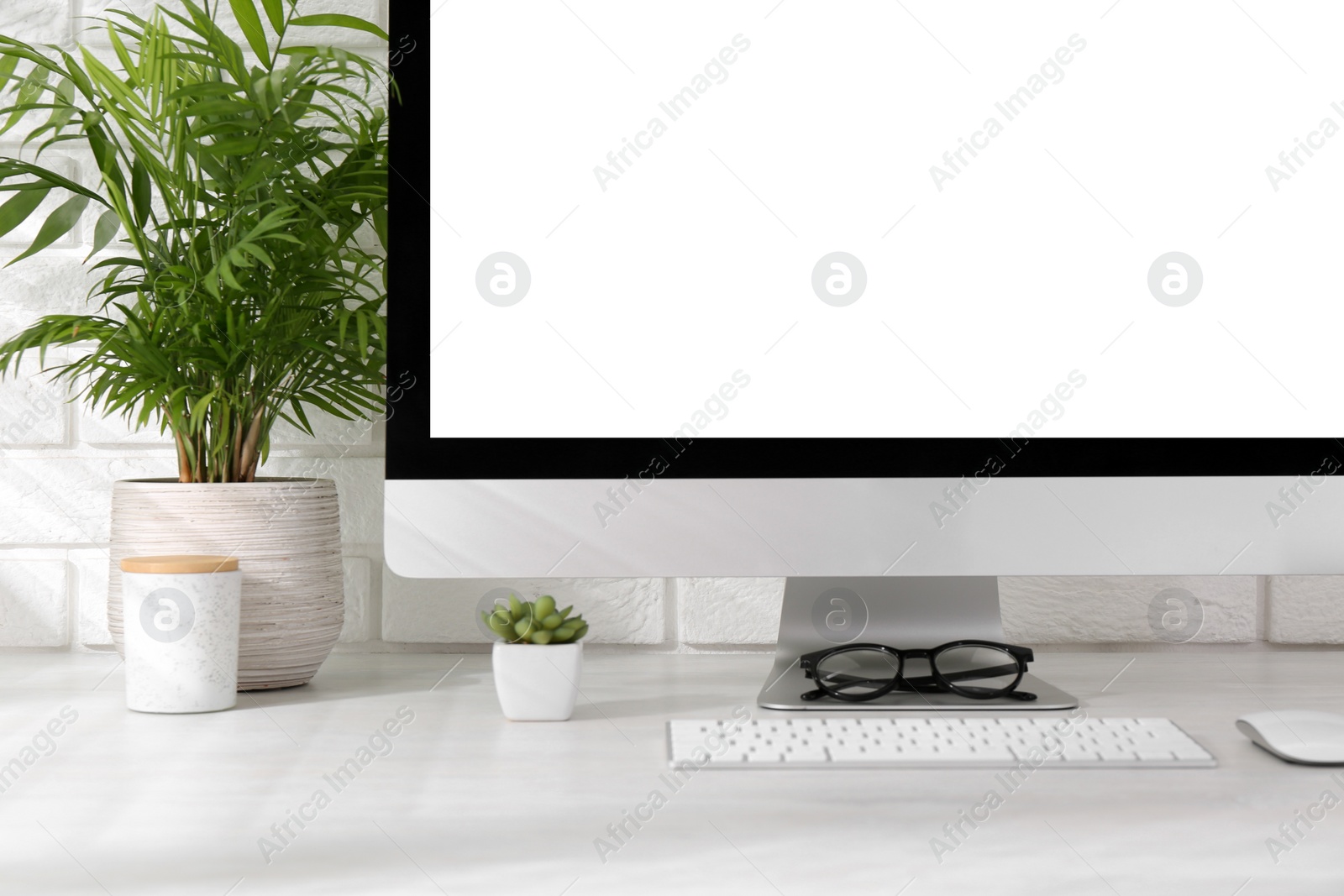 Photo of Office workplace with computer, glasses and houseplants on light table near white brick wall