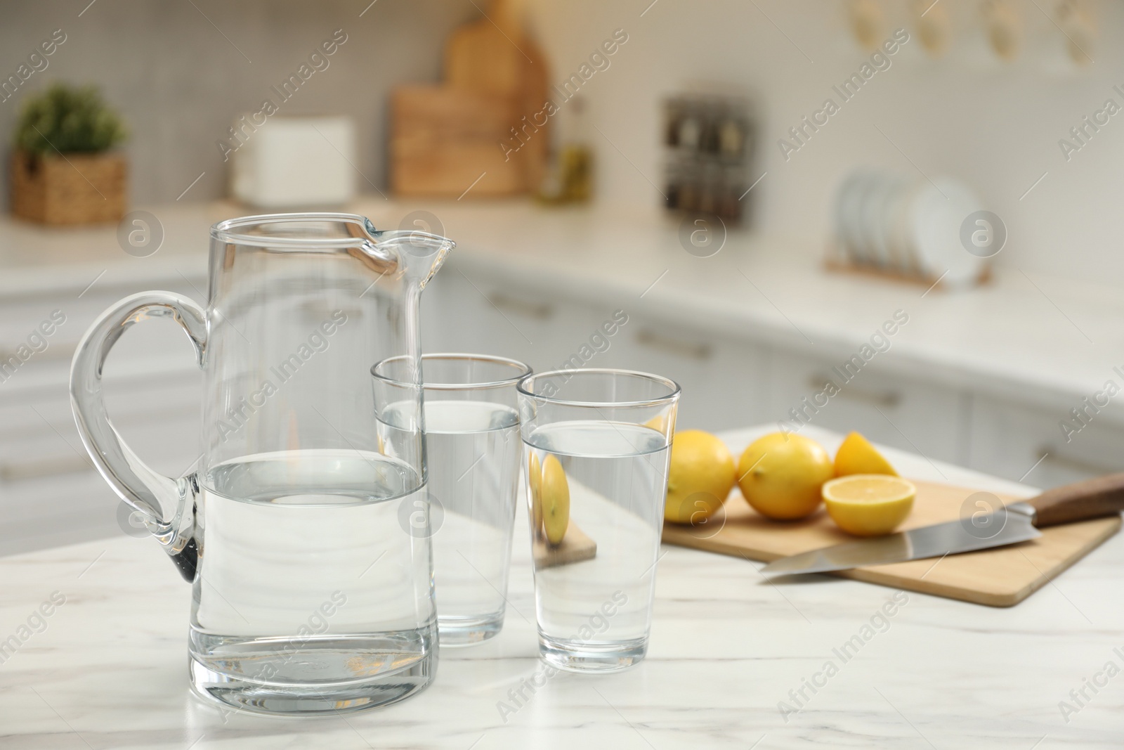 Photo of Jug, glasses with clear water and lemons on white table in kitchen