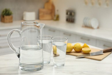 Jug, glasses with clear water and lemons on white table in kitchen