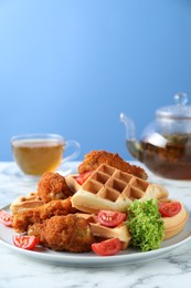 Photo of Tasty Belgian waffles served with fried chicken, tomatoes and lettuce on white marble table against light blue background, closeup. Space for text